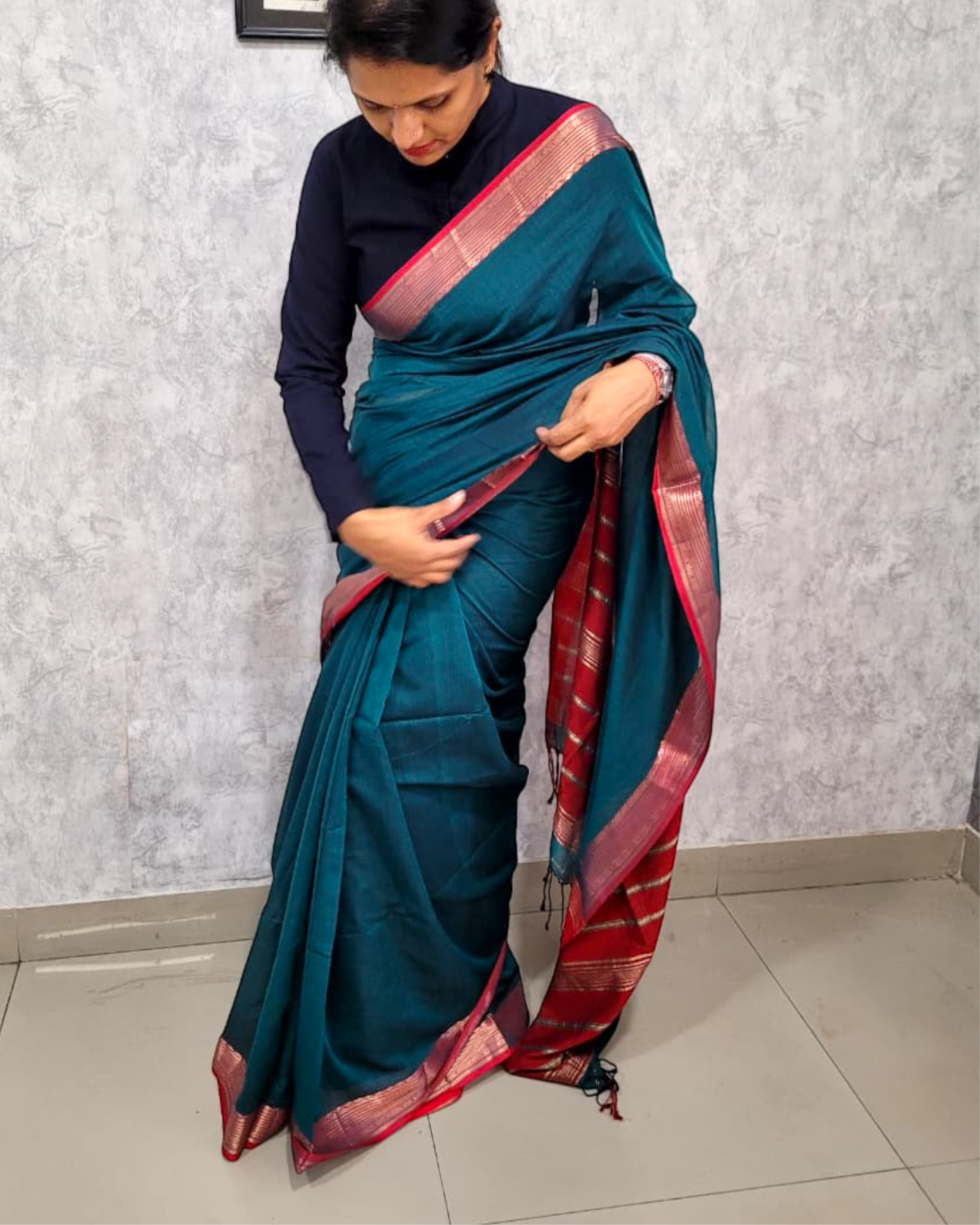Woman wearing a teal saree with a red border against a light gray wall.
